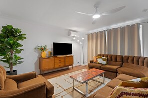 Ein modernes Wohnzimmer mit einer Sofagarnitur, einem Fernseher auf einem Holzkabinett, einer Topfpflanze und einem Deckenventilator.