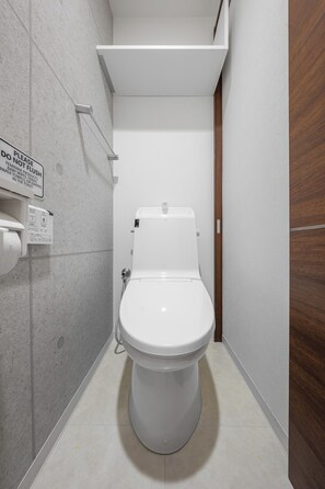 A compact, modern bathroom with a white toilet, a wall-mounted toilet paper dispenser, and a sign instructing to flush after use. - GRANDIA 202 (Sapporo)