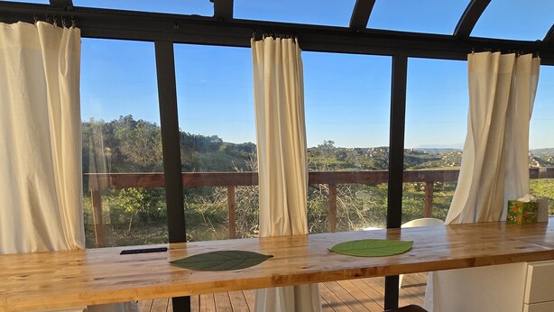 A room with a wooden table, two stools, a view of a forest through large windows, and a wooden ceiling structure. - Cozy Luxury Cabin in the De Luz Heights (Fallbrook)