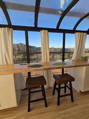 Ein Zimmer mit Holztisch, zwei Hocker, Blick auf einen Wald durch große Fenster und einer Holzbalkendecke.