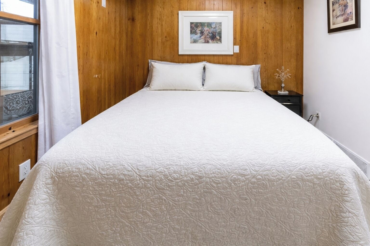 A bedroom with a wooden headboard, a large bed with white bedding, a framed picture on the wall, and a small bedside table with a vase.