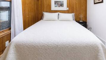 A bedroom with a wooden headboard, a large bed with white bedding, a framed picture on the wall, and a small bedside table with a vase.