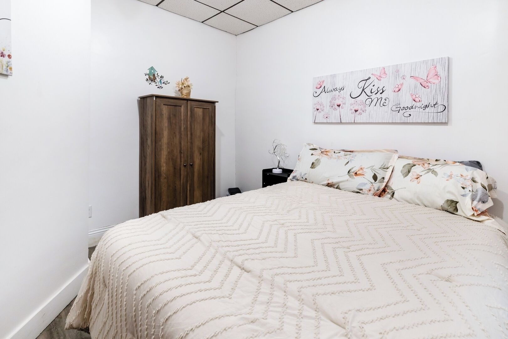 Une chambre à coucher comprenant un lit, une armoire en bois, une table de chevet, une décoration murale à motifs floraux et un luminaire au plafond.