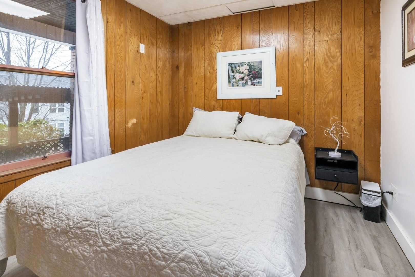 A bedroom with a wooden paneled wall, a bed with a white quilt, a framed picture above the bed, and a small bedside table with a vase.