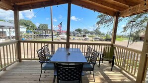 Eine überdachte Terrasse im Freien mit einem Holztisch und -stühlen, im Hintergrund Blick auf Boote und eine Flagge.