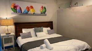 Una cama bien hecha con una colcha blanca y dos almohadas, una lámpara de noche y una obra de arte en la pared con pájaros coloridos.