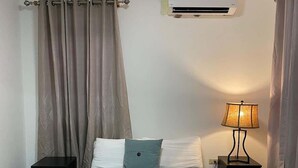 A hotel room with a single bed, a bedside table, a lamp, an air conditioning unit, and curtains. - Condado Beach Stay 1BR AC Wi-Fi Handicap (Condado)
