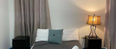 A hotel room with a single bed, a bedside table, a lamp, an air conditioning unit, and curtains.