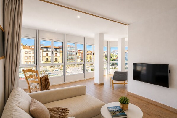 A modern living room with a sofa, a chair, a TV, and large windows offering city views. - Llanes Downtown Port View Apartment - Vega 2 (Llanes)