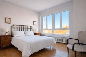 Ein Schlafzimmer mit einem großen Bett, Holznachttischen, einem Sessel und einem großen Fenster mit Blick auf das Dach.