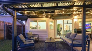 En udendørs patio med et overdækket siddeområde, herunder to sofaer og to stole, et lille bord og en potteplante.