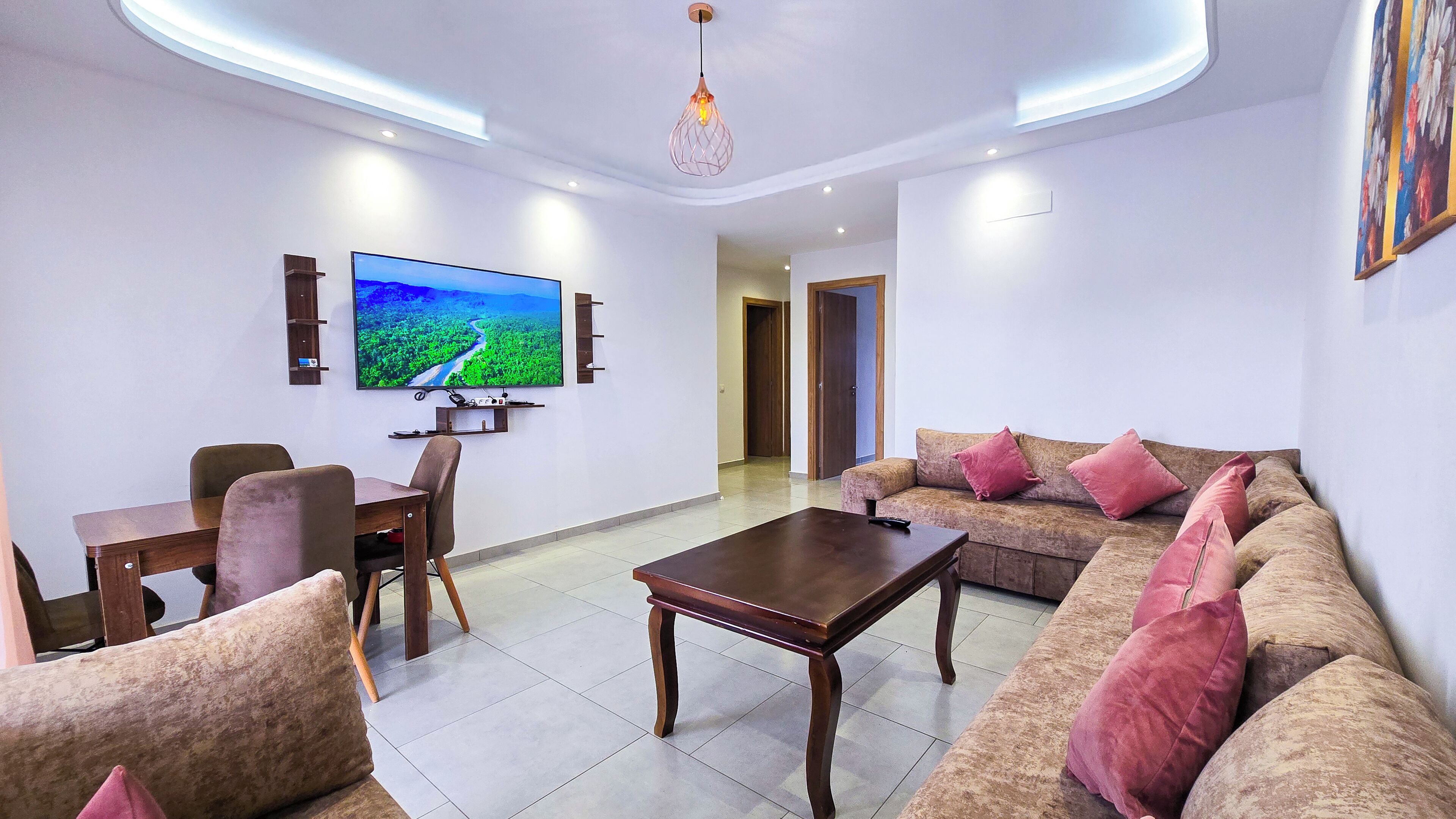 A modern living room with a flat-screen TV, a wooden coffee table, a sofa with pink cushions, and a dining area with chairs.