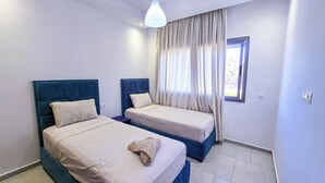 A hotel room with two beds, a window with curtains, a ceiling light, and a small window with a view of the outside.
