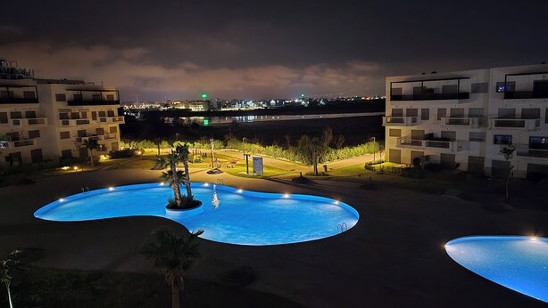A nighttime view of a pool area with two pools, surrounded by modern buildings and a distant cityscape.