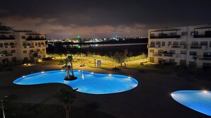 Szenischer Nachtblick auf einen Poolbereich mit zwei Pools, umgeben von modernen Gebäuden und einer weiter entfernten Stadtlandschaft.