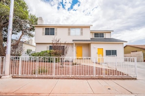 A two-story residential building with a yellow door and windows, surrounded by a white metal fence.