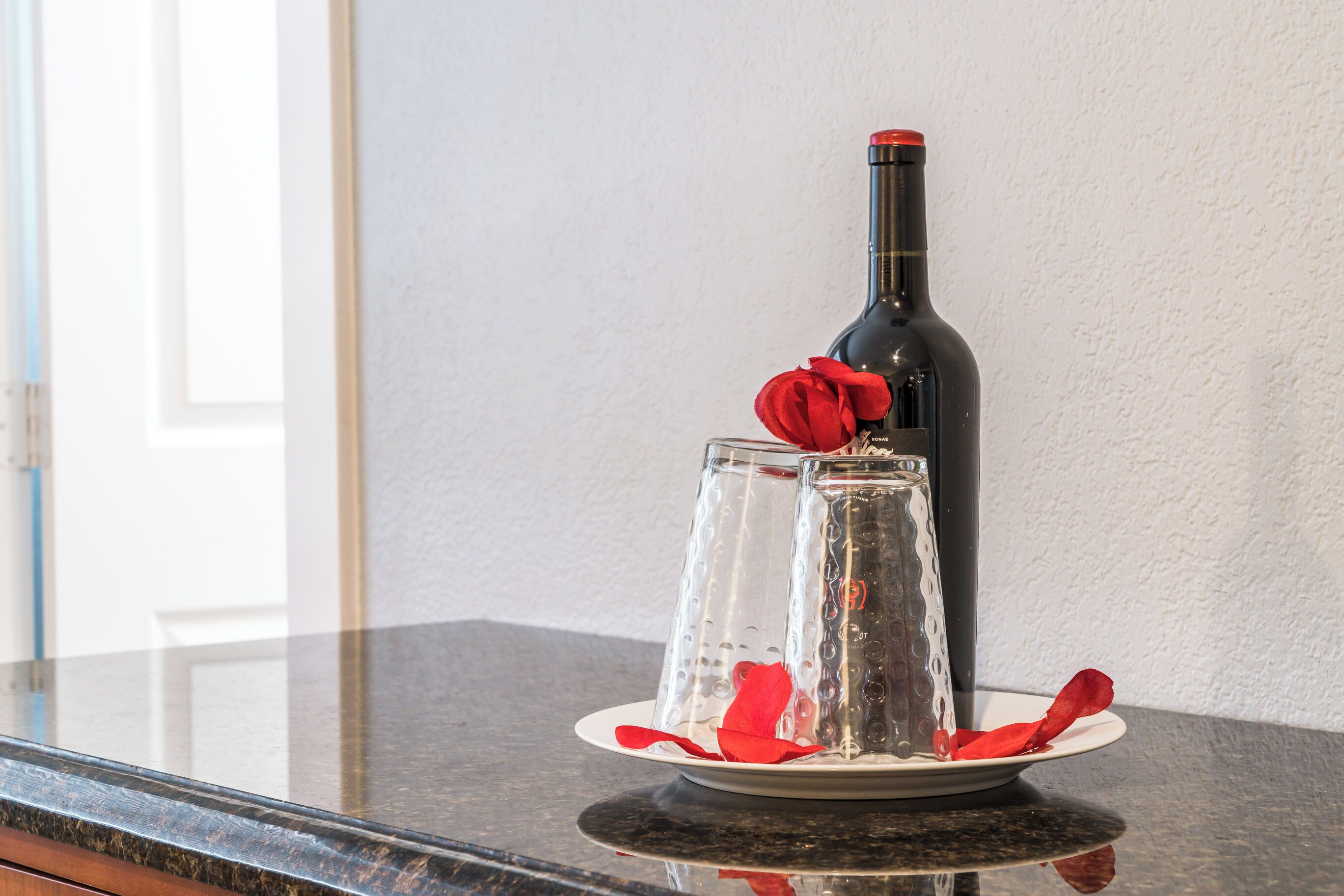 A wine bottle on a glass table with two glasses and red rose petals.