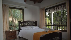 Une chambre à coucher avec un lit, une table de chevet, une fenêtre avec des barreaux en métal et une vue sur la verdure à l’extérieur.