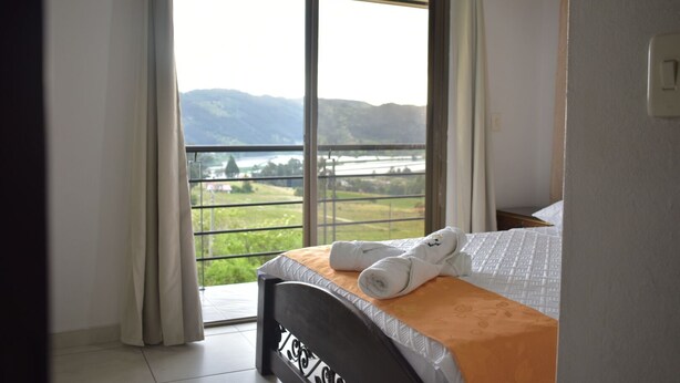Une chambre d’hôtel avec un lit, une fenêtre donnant sur une rivière et des montagnes, et un balcon avec une balustrade.