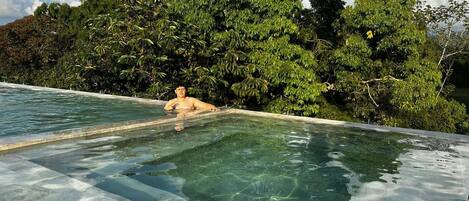 A man relaxing in a clear infinity pool with a view of dense foliage and a clear sky.