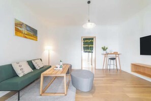 A modern living room with a green sofa, wooden coffee table, and a grey ottoman. There is a dining area with a small table and chairs. A large painting and a framed picture adorn the walls.
