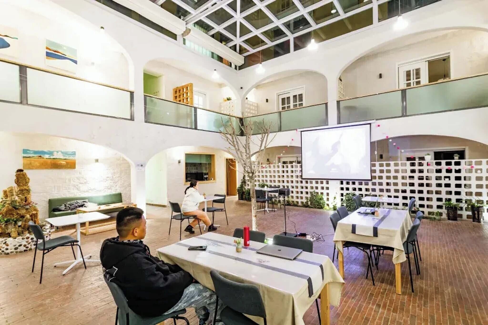 A spacious indoor area with multiple tables and chairs, a large screen for presentations, and a balcony with railings.