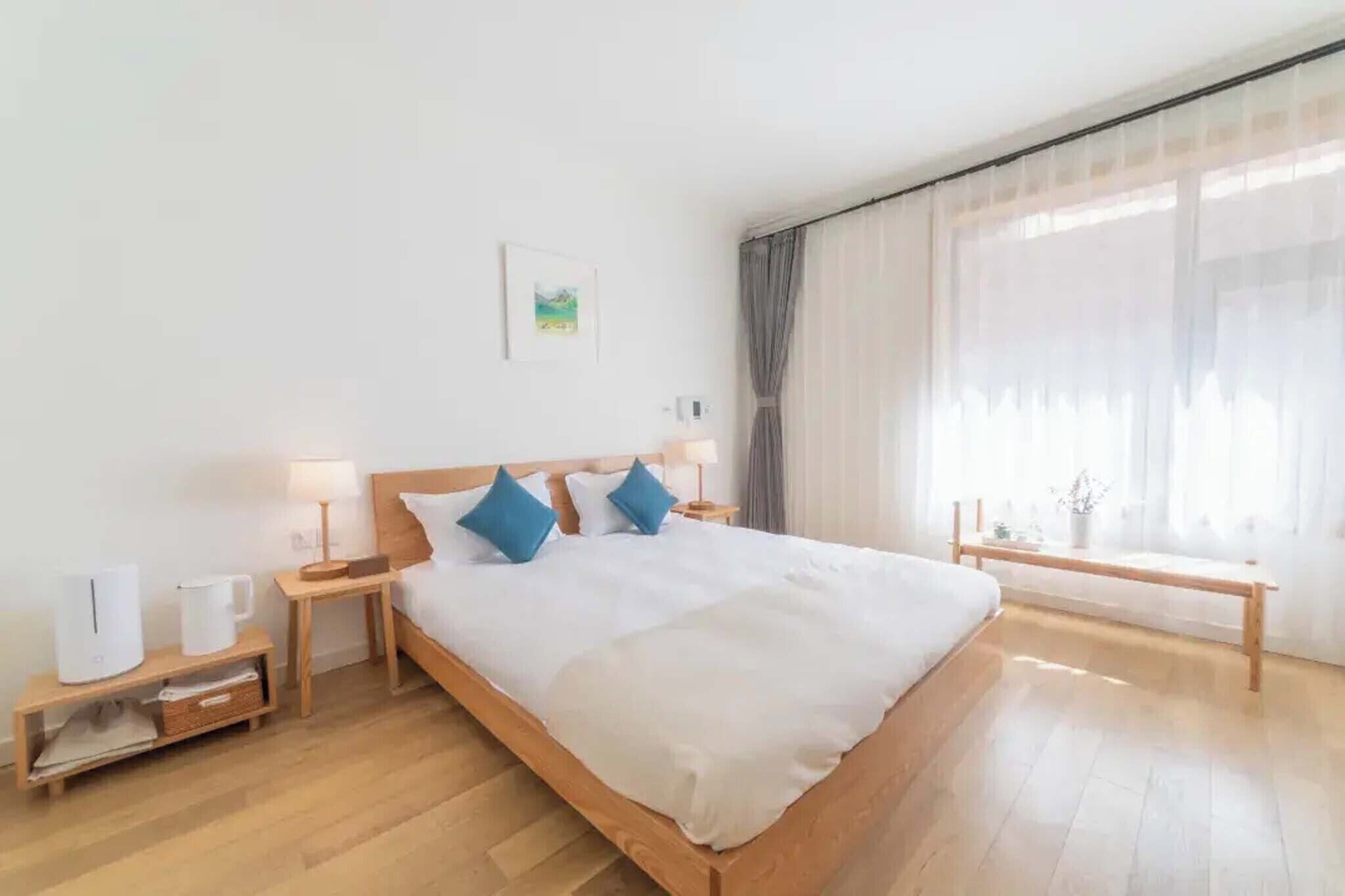 Una habitación de hotel moderna con una cama de madera, ropa de cama blanca, una mesita de noche con lámpara y otra mesita más pequeña con cafetera, y un ventanal con cortinas translúcidas.