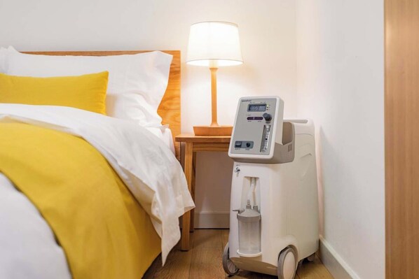 A hotel room with a bed, a bedside table with a lamp, and a medical device on the floor.