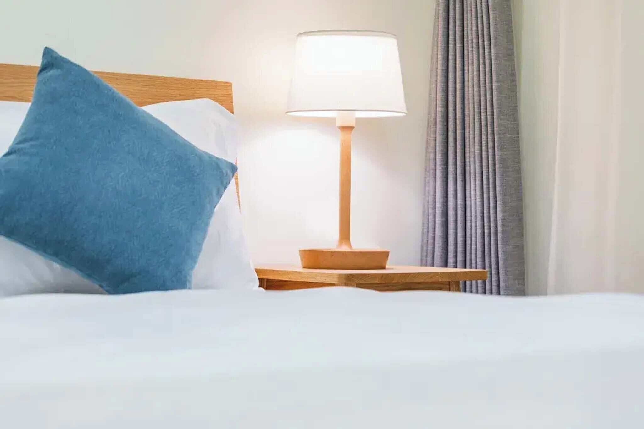 A neatly made bed with a blue pillow, a wooden nightstand with a lamp, and grey curtains.
