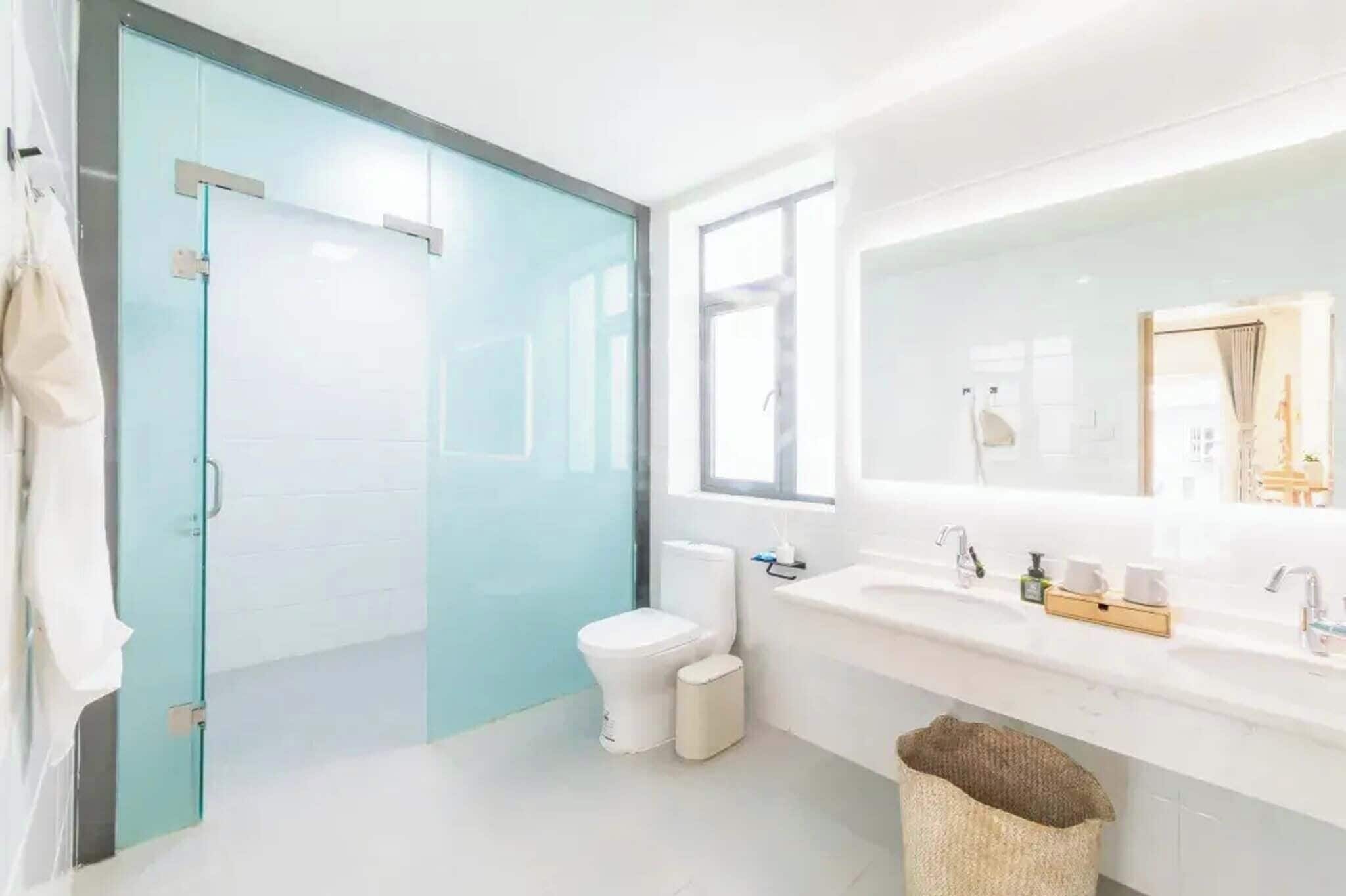 A modern bathroom with a glass shower enclosure, a white toilet, a sink with a large mirror, and a window providing natural light.