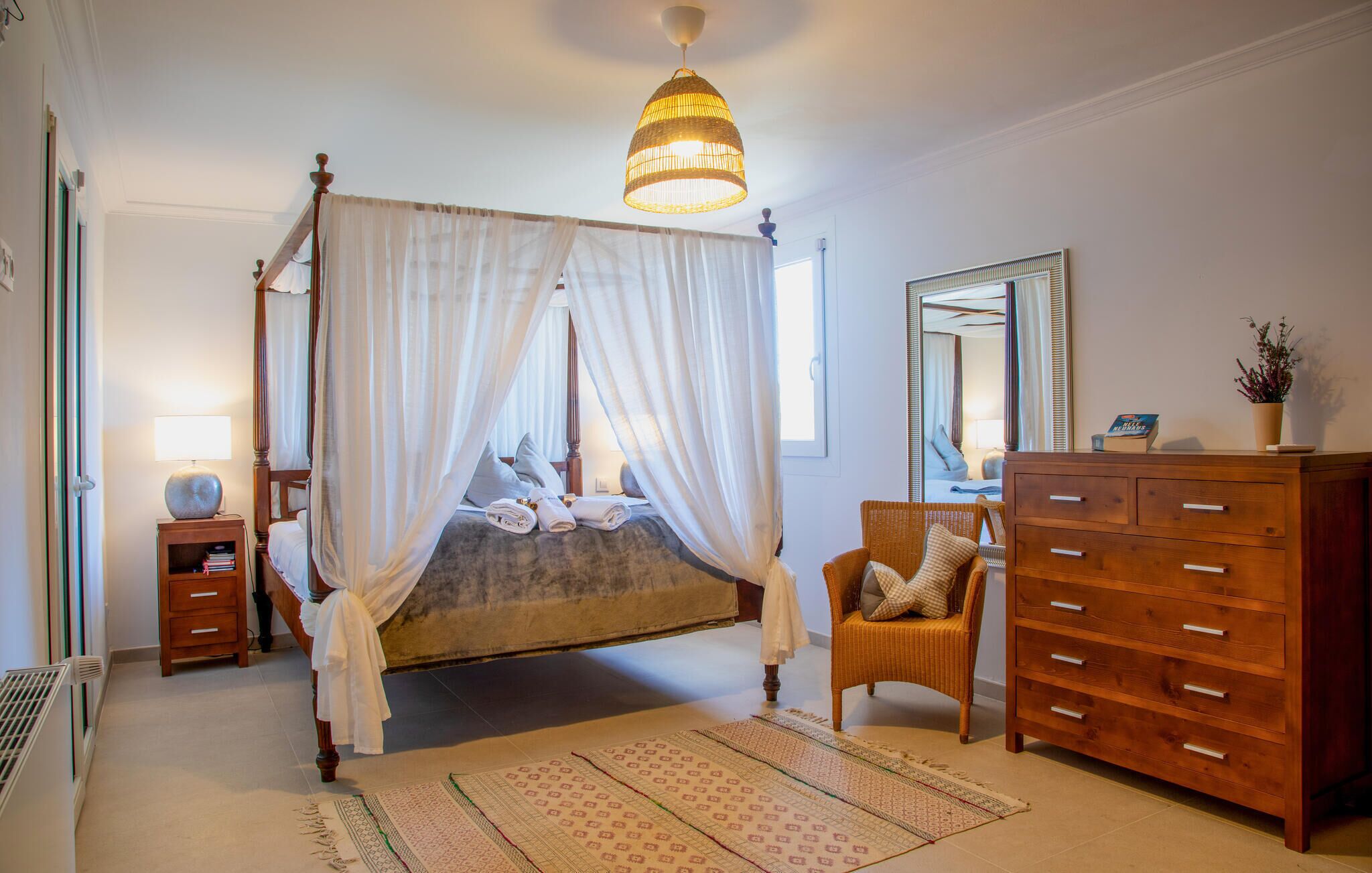 A four-poster bed with a canopy, a wooden dresser, a wicker chair, and a large mirror.