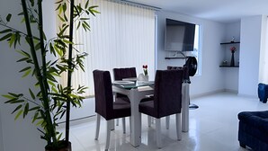 Un espace repas moderne avec une table blanche et des chaises foncées, une grande fenêtre avec des stores, un téléviseur à écran plat fixé au mur et une plante en pot.