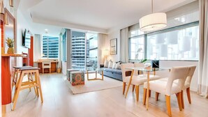 Ein modernes Hotelzimmer mit Essbereich, einer Couch, einem Fernseher und einem großen Fenster mit Blick auf die Stadt.