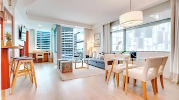 Quarto de hotel moderno com área de refeições, sofá, TV e uma grande janela com vista para a cidade.