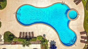En bild tagen från luften som visar en pool med en ovanlig form, där en del är cirkelformad. Poolen omges av solstolar och en liten trädgårdsdel.