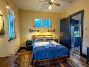 A bedroom with a bed, bedside tables, a ceiling fan, a window, and a door leading to a bathroom.