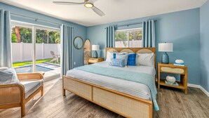 Quarto com uma cama grande, duas mesas de cabeceira com luminárias, um ventilador de teto e vista para um quintal com piscina e cadeiras.