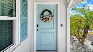 Uma porta azul clara com uma grinalda e a palavra 'Beach', uma janela com persianas e uma palmeira do lado de fora.