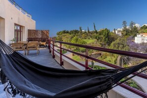 A balcony with a hammock, wooden furniture, and a view of a landscaped garden and buildings. - Stay In St. Catarina Park, by PAUSA H. Rentals (Funchal)