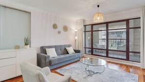 Ein modernes Wohnzimmer mit einem grauen Sofa, einem Glastisch und einem großen Fenster mit Blick auf Gebäude.