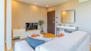 Ein modernes Hotelzimmer mit einer Couch, einem kleinen Tisch mit Schale und Pflanze, einem Fernseher auf einem Schrank, einem Spiegel und einer Tür.