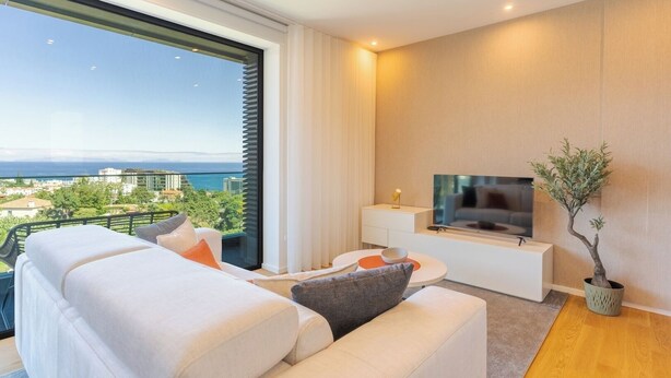 Ein modernes Wohnzimmer mit Sofa, Fernseher und Blick auf Stadt und Meer.