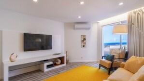Un moderno salón con un televisor de pantalla plana, un sofá, una alfombra amarilla y vista al mar a través de amplios ventanales.