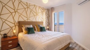 Un dormitorio moderno con un papel tapiz de diseño geométrico, una cama grande con ropa de cama blanca y vistas al mar a través de un ventanal corredero.