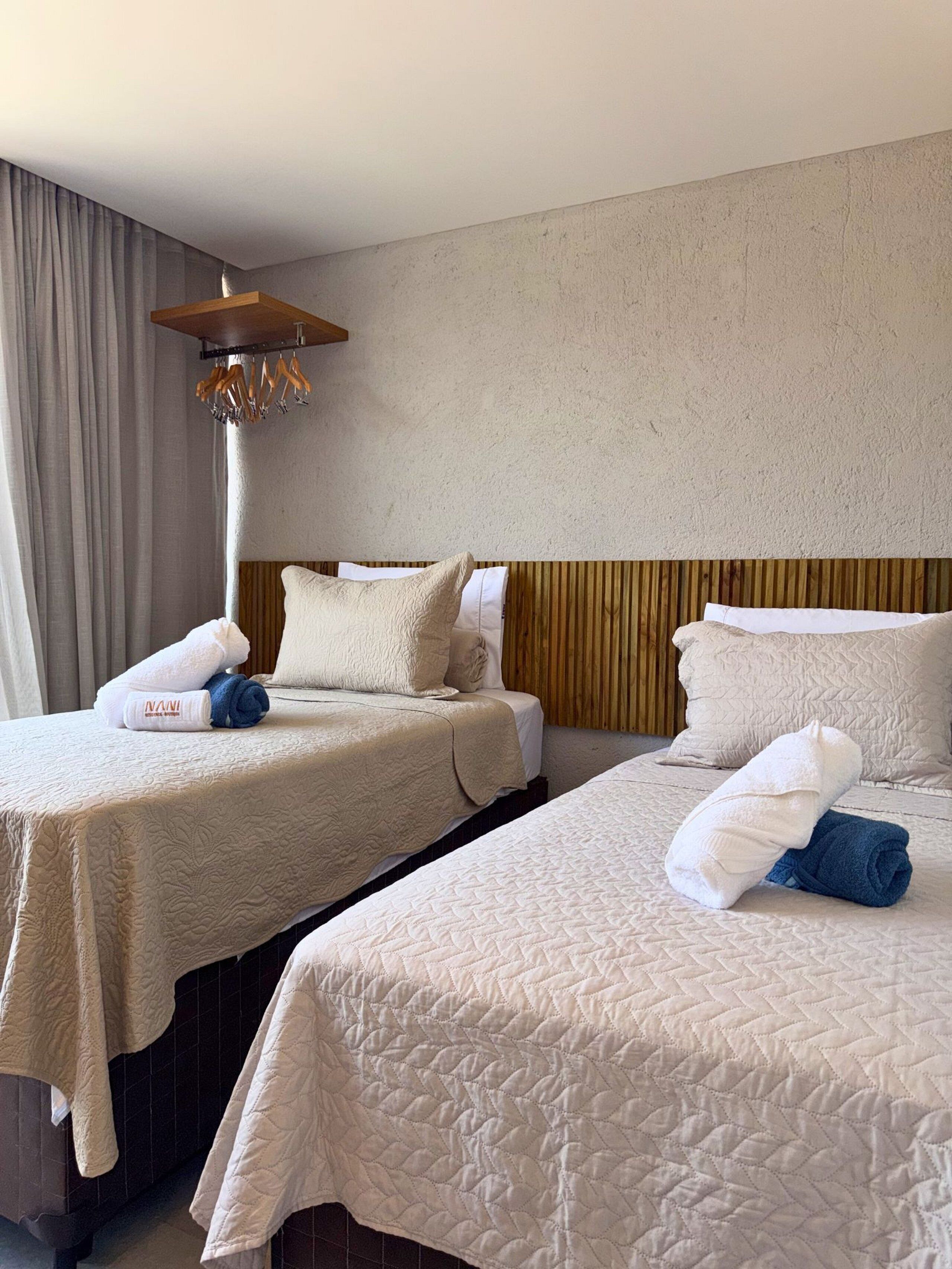 Ein Hotelzimmer mit zwei Betten, einem Holzkopfstück, einer Hängelampe und einem großen Fenster mit Vorhängen.