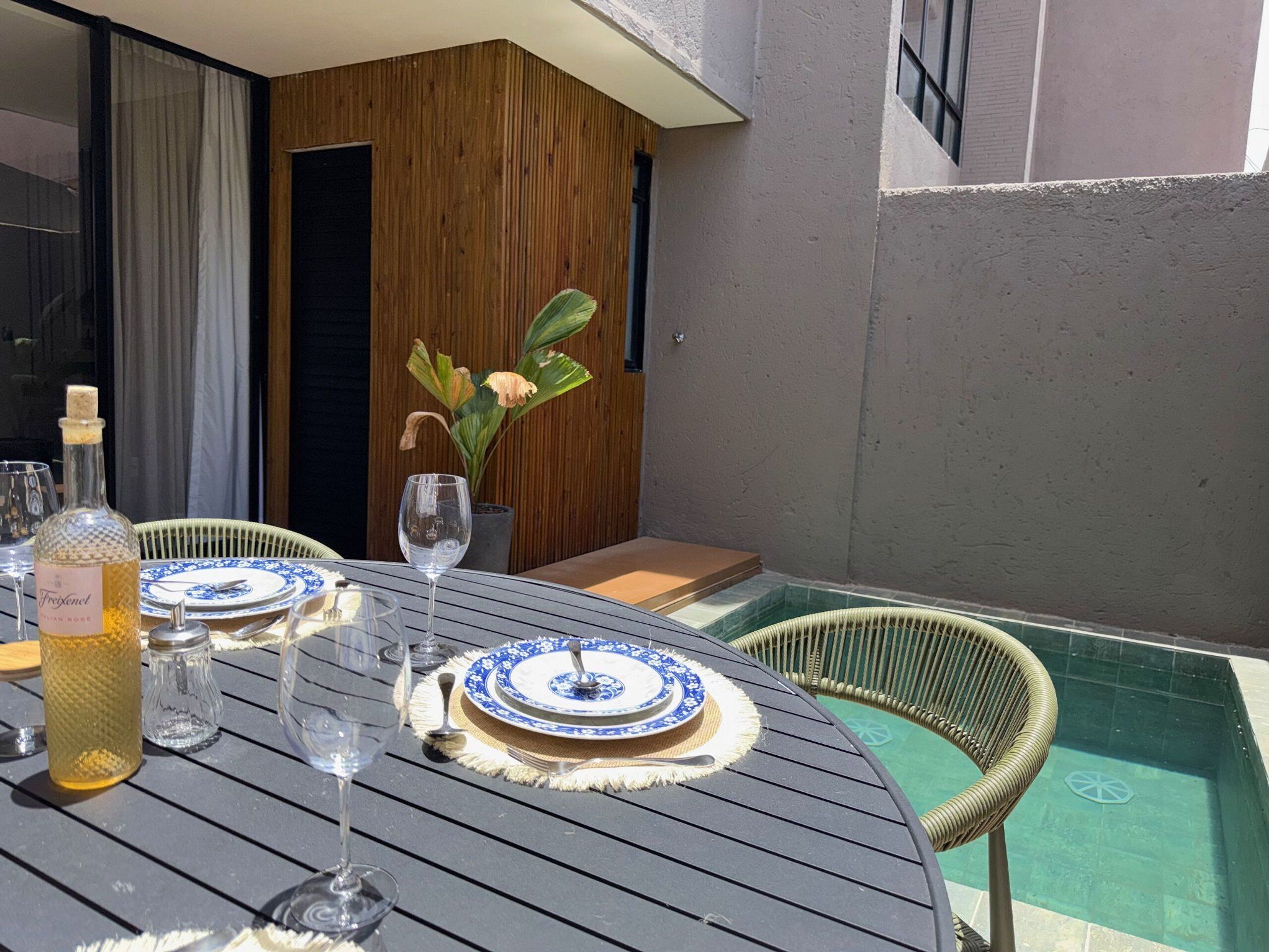 Una mesa de comedor con platos, vasos y una botella, rodeada de sillas de mimbre y una pequeña planta en maceta.