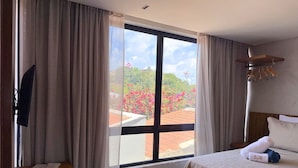 A hotel room with a bed, a desk, a window with curtains, and a view of a garden. - MHV Milagres - Nani Villas Boutique (São Miguel dos Milagres)
