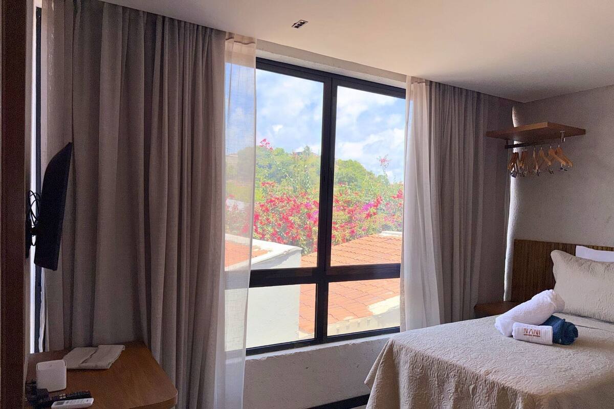 Ein Hotelzimmer mit einem Bett, einem Schreibtisch, einem fenster mit Vorhängen und Blick auf einen Garten.
