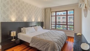 Ein Hotelzimmer mit einem großen Bett, einem Nachttisch, einer Lampe, einem Fenster mit Blick auf Gebäude und einem Wandbild.