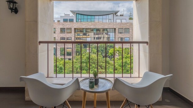 Ein Balkon mit zwei weißen Stühlen und einem kleinen runden Tisch, mit Blick auf ein modernes Gebäude mit großen Fenstern und Grünflächen.